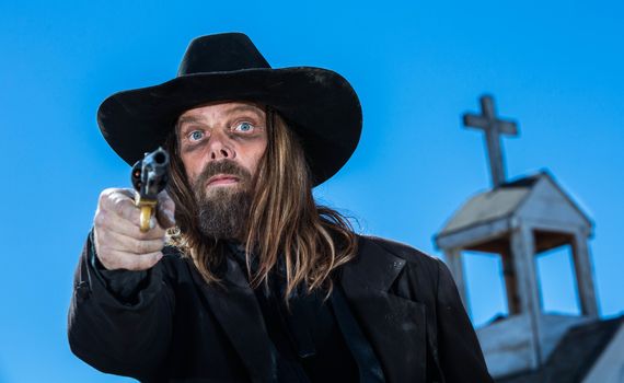 A serious cowboy holding a gun near a church