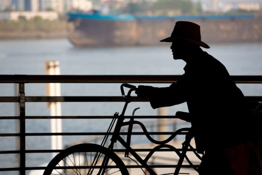 Silhouette of a Chinese man pushing his bicycle with the river and a boat in the background in shanghai, china 2013.