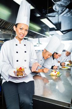 Asian Indonesian chef along with other cooks in restaurant or hotel kitchen cooking, finishing dish or plate for dessert