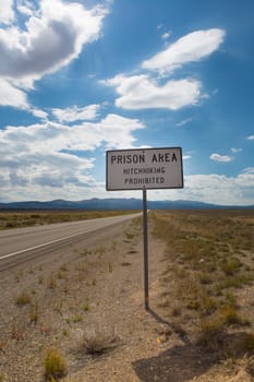 Sign reading "Prison area, hitchhiking prohibited" stands in front of a prison/penitentiary in the desert landscape beneath a cloud filled blue sky. Taken in the Utah state.