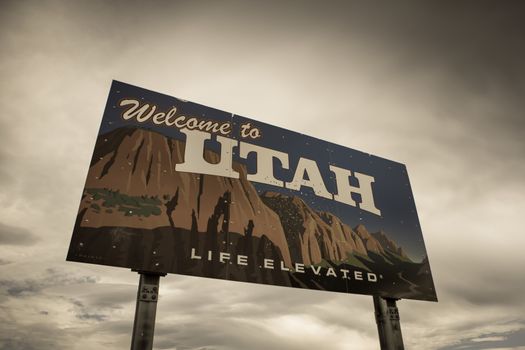 On the border of Nevada and Utah, a Welcome to Utah sign marks the separation of the two states underneath a cloud filled gray sky. Retro style image