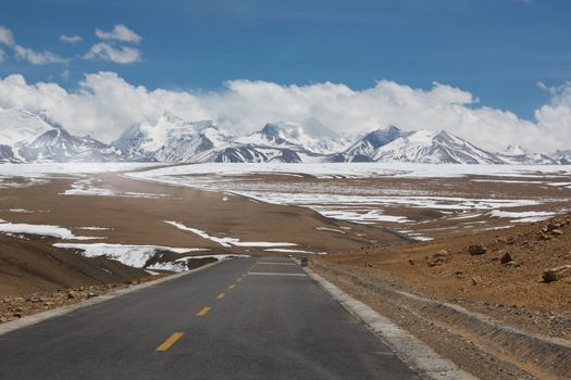 Straight road with a yellow dotted line in the middle with Tibetan landscape of Mountains in the background. The road seems to go toward the top of the snowed mountain in the cloud.