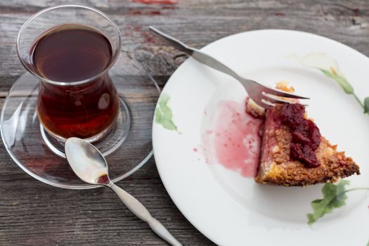 Slice of delicious cheesecake topped with a berry compote and served with a tall glass of freshly brewed Turkish tea on an old wooden table
