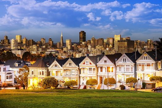 The Painted Ladies of San Francisco, California sit glowing amid the backdrop of a sunset and skyscrapers. 