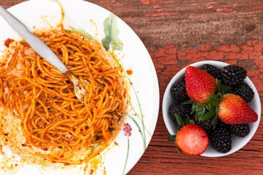 Overhead view of a messy plate of spaghetti Bolognese in a rich tomato sauce and fresh berry dessert with strawberries and blackberries on a grungy old wooden table with peeling paint
