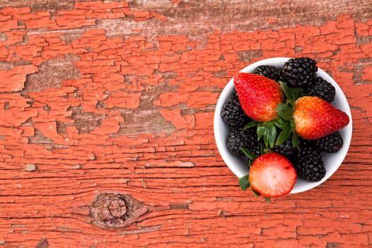 View from above of a small bowl of fresh ripe mixed berries containing strawberries and blackberries on a rustic grungy old wooden picnic table with peeling red paint and copyspace for your text