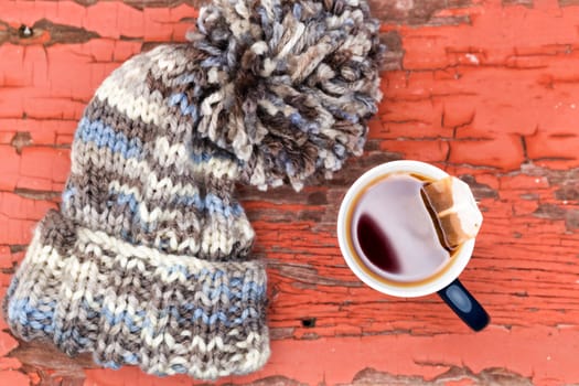 View from above of a cosy knitted woolly winter hat with a large pompom beside a cup of fresh hot tea on a grungy wooden surface with peeling red paint