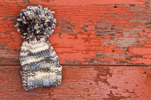 Cute warm woolen winter hat with a large pompom lying on an old wooden picnic table with a grungy surface of peeling red paint with copyspace for your text