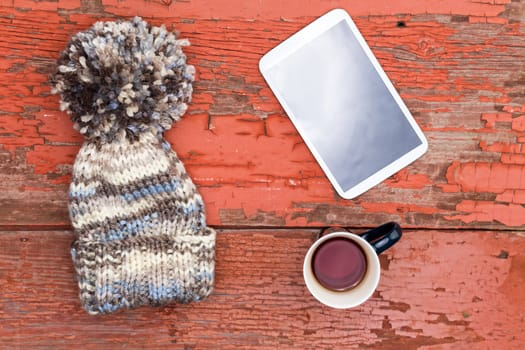 Woolly winter cap with a large decorative pompom lying with a tablet computer and mug of fresh tea on a grungy wooden table with peeling paint