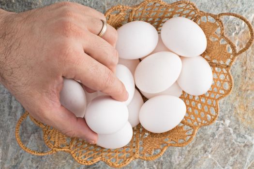 Hand of a man wearing a wedding ring taking a traditional Easter egg from an ornamental woven wicker Easter basket filled with fresh white eggs