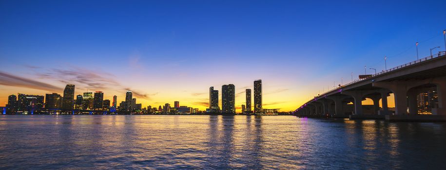 Miami city skyline panorama at dusk with urban skyscrapers and bridge over sea with reflection 