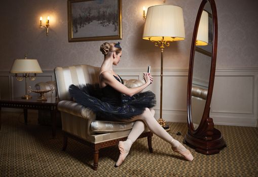 Ballerina in black tutu sitting in armchair in front of mirror and taking self portrait with camera on her cell phone