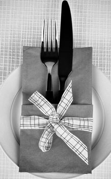 Elegant Table Setting with Fork and Table Knife into Napkin Decorated with Checkered Bow on Plate. Monochrome View