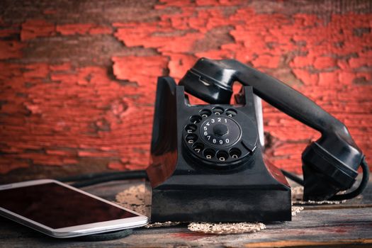 Old rotary telephone with its receiver off the hook alongside a tablet computer showing the old-fashioned and modern forms of communication against a grungy wooden wall with peeling paint