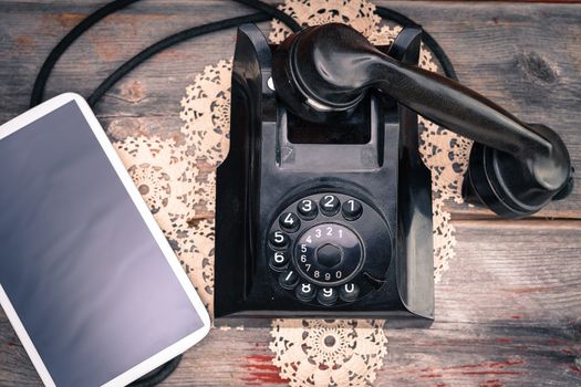 Tablet computer with a blank screen lying alongside a retro rotary telephone with its handset off the hook on a decorative doily, high angle view