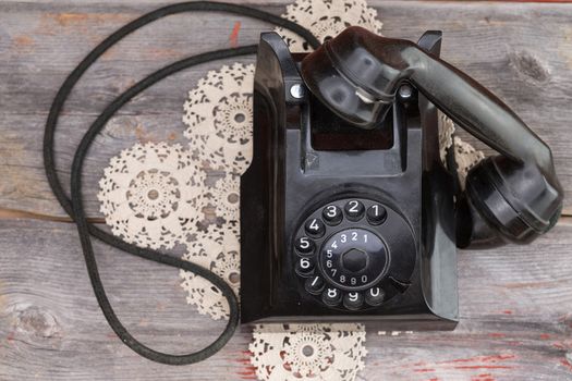 Old rotary telephone with the handset off the hook standing on a decorative patterned crocheted doily on a rustic weathered wooden table