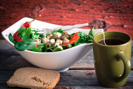 Bowl of delicious fresh baby spinach, tomato and feta salad with a mug of hot tea and a slice of bread served for lunch on a rustic wooden background with peeling paint