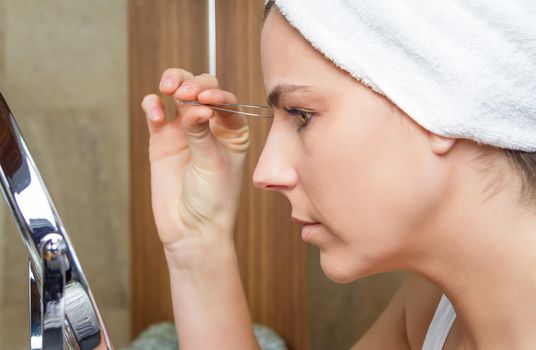 beautiful young woman plucking her eyebrows with tweezers in front of mirror on the bathroom