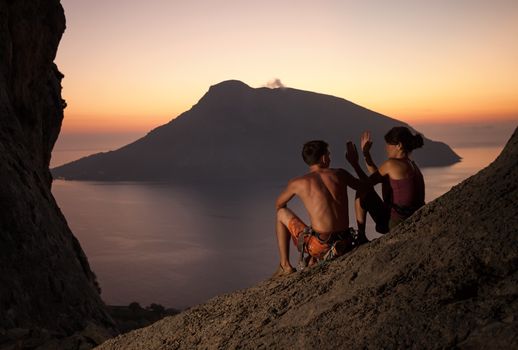 Two rock climbers having rest at sunset. Climbers giving high five and cheering. With picturesque view of Telendos Island in front. Kalymnos Island, Greece.
