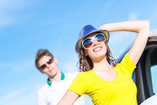 young attractive woman in sunglasses and hat stands next to a car, holds her hat and looks at the sun