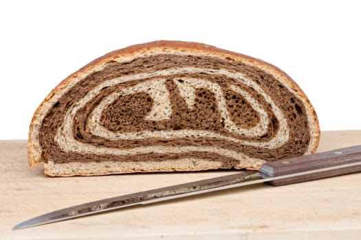 A halved loaf of freshly baked bi-colour gourmet bread with a distinctive spiral brown and white pattern with a bread knife on a wooden board over a white background