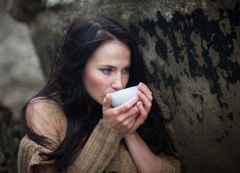 Young cute girl in a knitted sweater against a background of an old concrete wall drinking tea from a white cup
