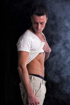 Handsome, fit young man pulling up t-shirt revealing abs. Dark background and smoke
