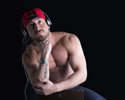 Muscular man sitting, listening to music on headphones, looking up, black background