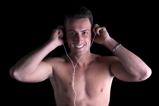 Athletic shirtless man listening to music on headphones, looking at camera smiling, black background
