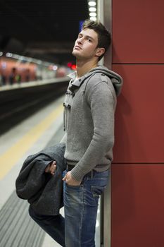 Handsome young man standing against wall in train or subway station, looking at camera