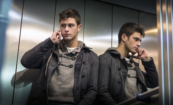 Handsome young man in elevator (lift) using cell phone, his reflection on mirror