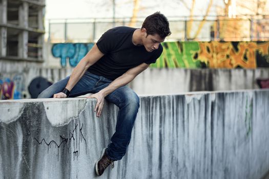Young man sitting and balancing on dirty wall, looking down, urban environment