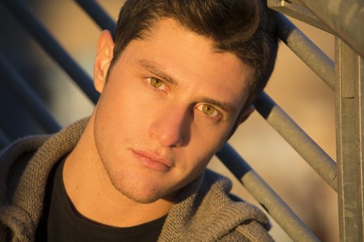 Headshot of handsome young man, lit by sunset light, leaning against handrail