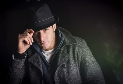 Attractive young man at night, wearing winter coat, tipping fedora hat while looking at camera