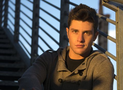 Headshot of handsome young man, lit by sunset light, leaning against handrail