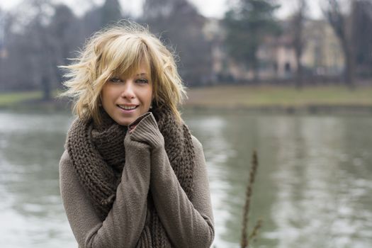 Pretty young blonde woman outdoors, wearing wool scarf and sweater near river or lake