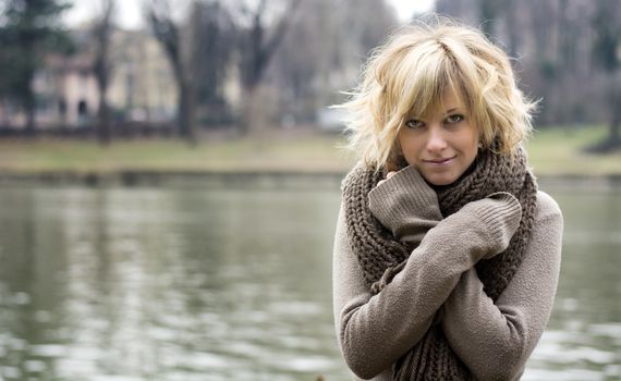 Pretty young blonde woman outdoors, wearing wool scarf and sweater near river or lake