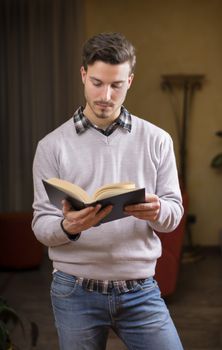 Handsome young man reading book at home in his living-room, standing