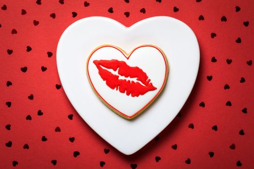 Valentine cookie in shape of heart on white plate. Red paper background with confetti. Valentine's Day composition. Top view 
