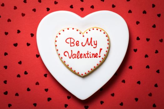 Valentine cookie in shape of heart on white plate. Be my valentine. Red paper background with confetti. Valentine's Day composition. Top view 