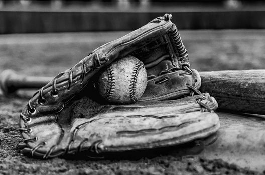 Baseball glory days with ball in glove and bat on base on field.  Monochrome image with outfield wall in background.