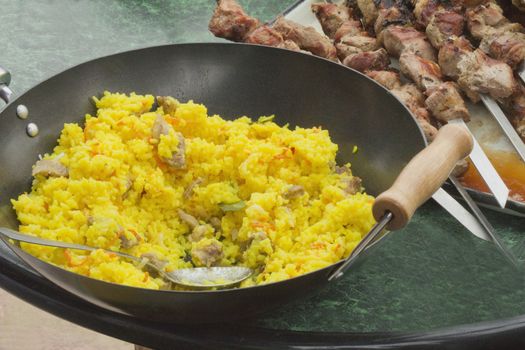 On the table are a large frying pan with a delicious plov and toasted shish kebab skewers