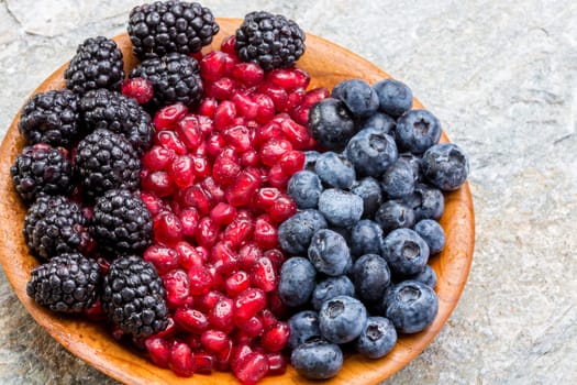 Wooden bowl of fresh blueberries, blackberries and pomegranate seeds arranged in colourful rows, high angle view on an old stone countertop