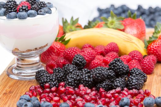 Preparing a delicious creamy fruit dessert with rows of colourful clean tropical fruit on a wooden counter including blueberries, blackberries, raspberries, strawberries, a banana and pomegranate