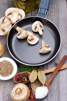 Preparing to Roast Edible Champignon Mushrooms in Frying Pan with Spices in Wooden Spoons  and Raw Ingredients closeup on Rustic Wooden background. Top View