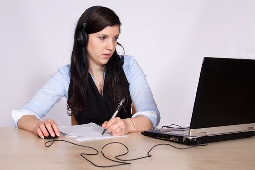 Young woman with headsets phone and writes with a pen on paper and sitting at table with laptop