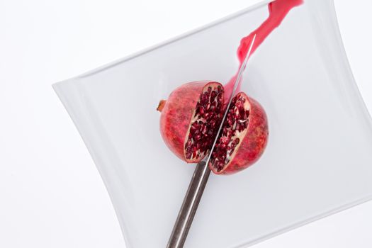 Fresh pomegrate being cut in half with a knife on a white plate with sweet juice running out as the pulpy seeds or arils or exposed, overhead view