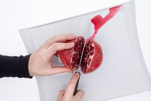 Clean hands of a man or chef cutting a fresh whole pomegranate in half exposing the ripw red pulpy seeds or arils and releasing fresh juice, overhead view on a white plate on a white background