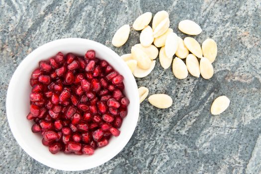 A white bowl of seeds of fresh pomegranate, next to peeled almonds, natural source of minerals and vitamins, on a stone counter top, high-angle close-up shot