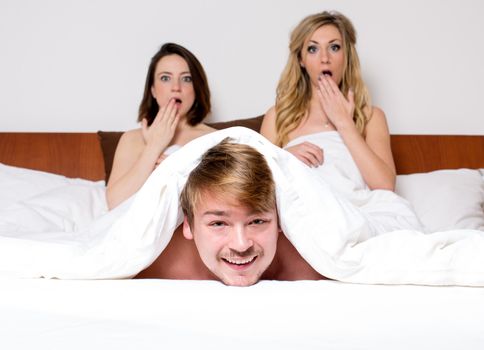Humorous conceptual image of a young man peeking out from under bedclothes with a cheeky grin on his face watched in horror by two young women sitting up at the other end of the bed